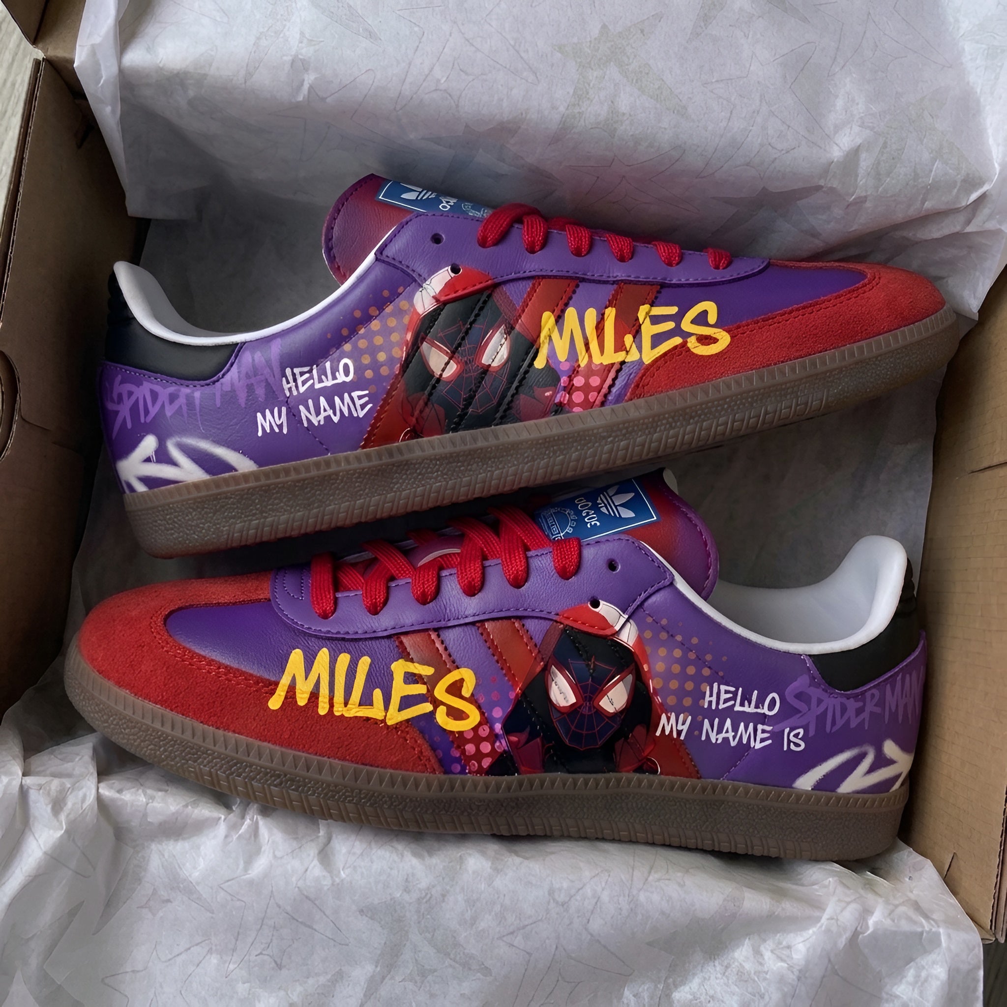 A pair of custom Spider-Man sneakers in a cardboard shoebox with white tissue paper. The shoes feature a purple and red color scheme with comic-style graphics of Miles Morales, yellow 'MILES' text, and white 'HELLO MY NAME IS' text.