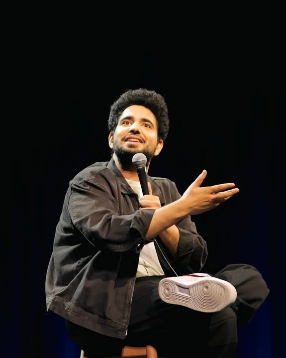 Samay Raina on stage with a microphone against a black background wearing knickgasm sneakers