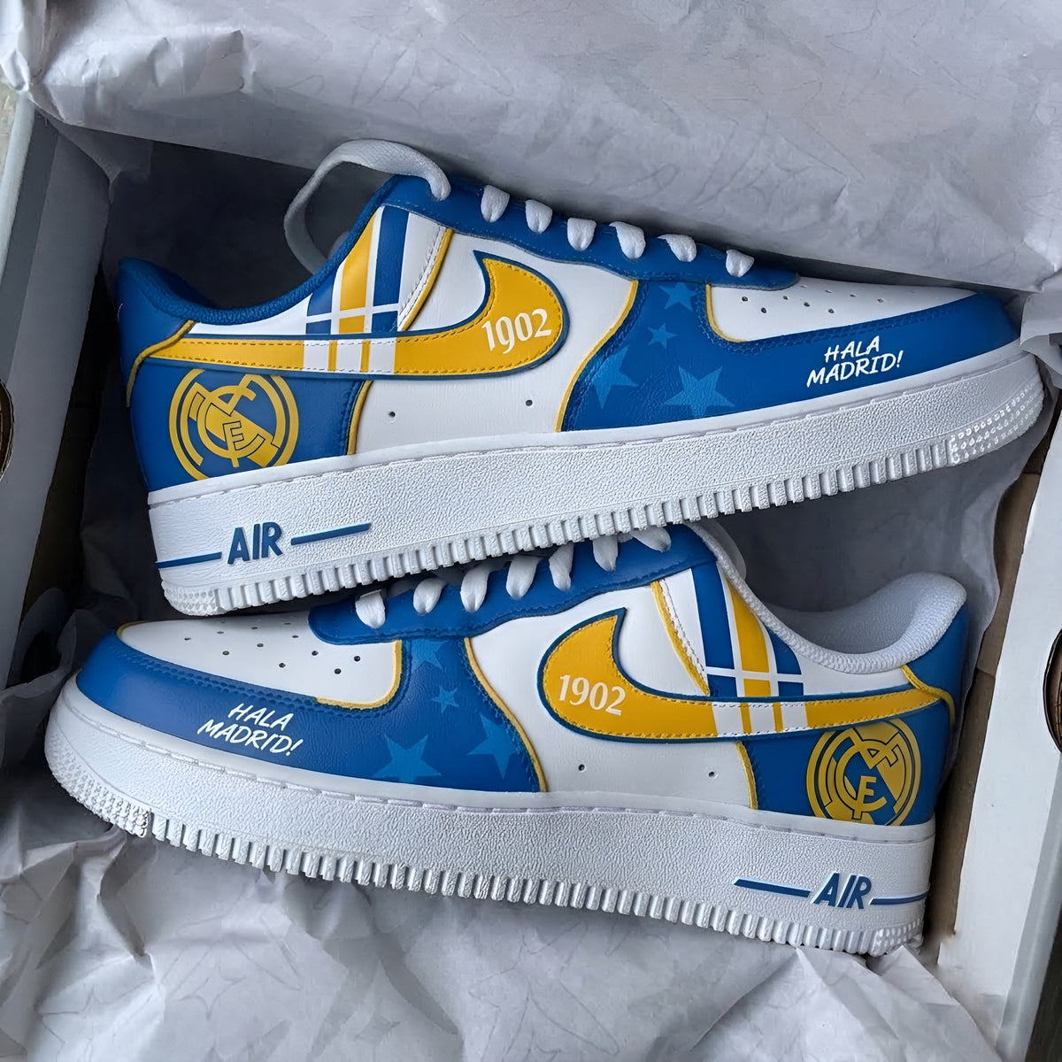 A pair of customized Real Madrid Nike Air Force 1 Low sneakers in blue, white, and yellow, resting inside a white shoebox lined with white tissue paper.